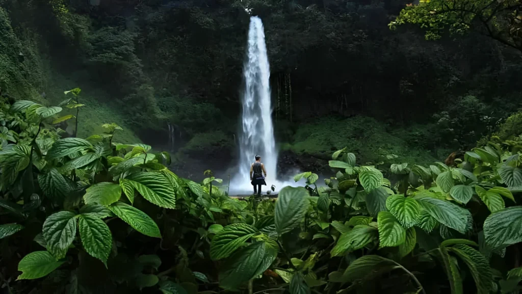 Serene and lush tropical scene, with a man standing in front of a majestic waterfall surrounded by dense greenery. The man is positioned centrally, facing away from the camera towards the waterfall, which cascades down a rocky cliffside into a misty veil.